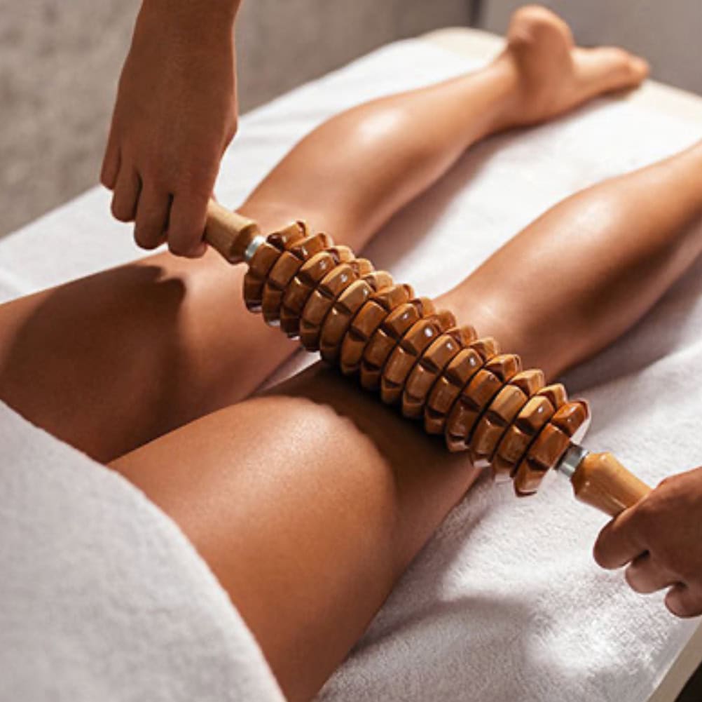 Person zeigt Massage mit Holzrolle an Beinen, entspannter Ausdruck, hellbraune Haut, weiße Abdeckung.